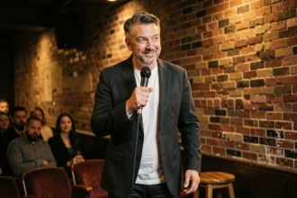 Comédien masculin sur scène dans un club de comédie tenant un micro avec un sourire complice face au public