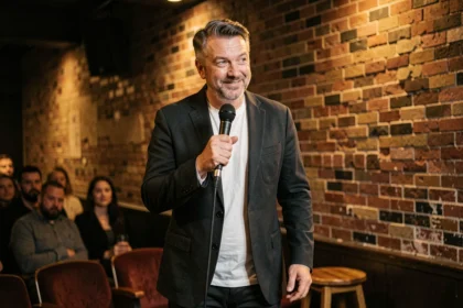 Comédien masculin sur scène dans un club de comédie tenant un micro avec un sourire complice face au public